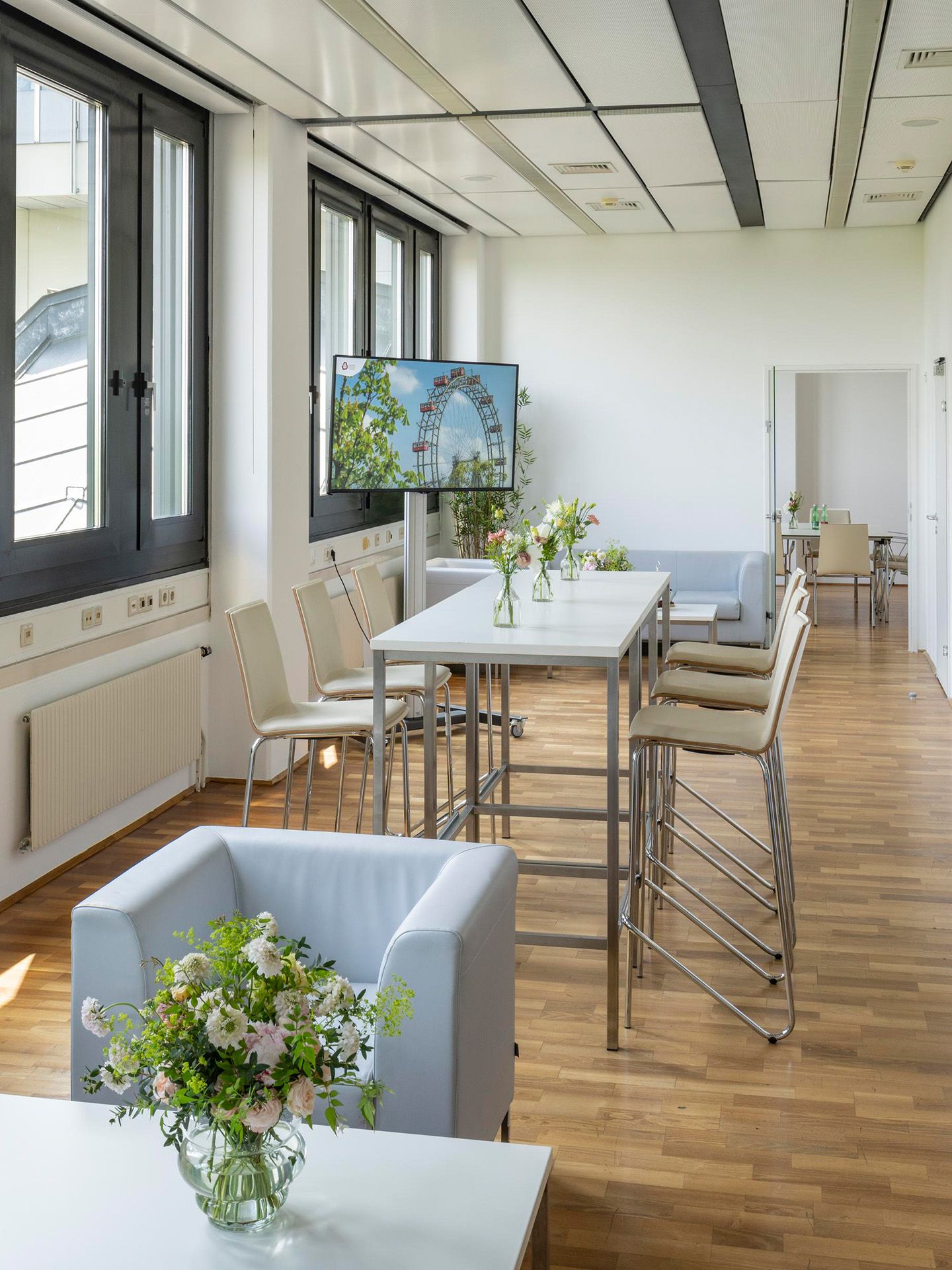 Foto: Raum mit Holzboden, hellen Moebeln, einem Fernseher und Blumen. Durch eine offene Tuer ist ein Nebenraum sichtbar. Durch die Fenster ist ein anderes Gebaeude sichtbar.