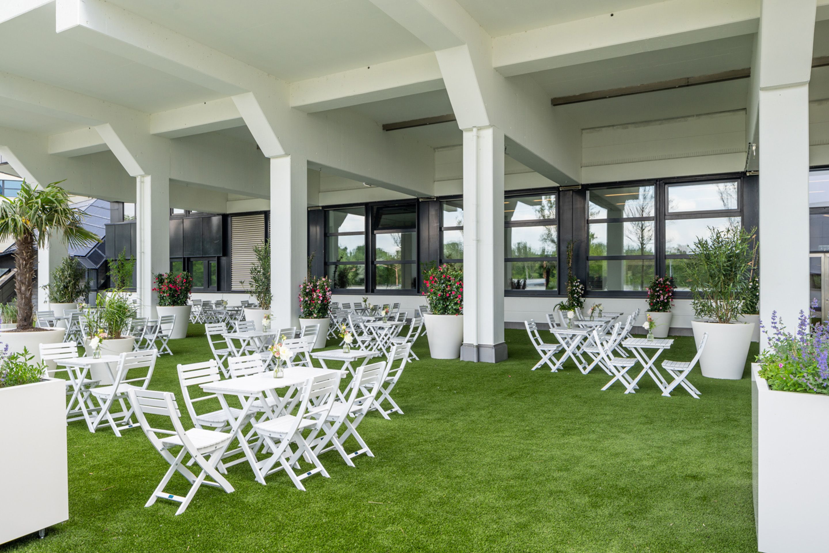 Foto: Sitzplaetze auf einem gruenem Teppich bei einem Gebaeude mit Saeulen, Blumentoepfe.