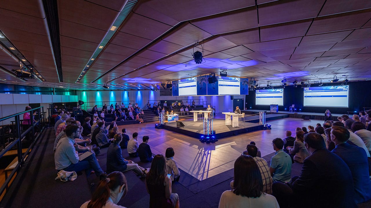 Foto: hell beleuchteter Podiumsdiskussions-Bereich mit einigen Panel-Teilnehmern vor einem Publikum