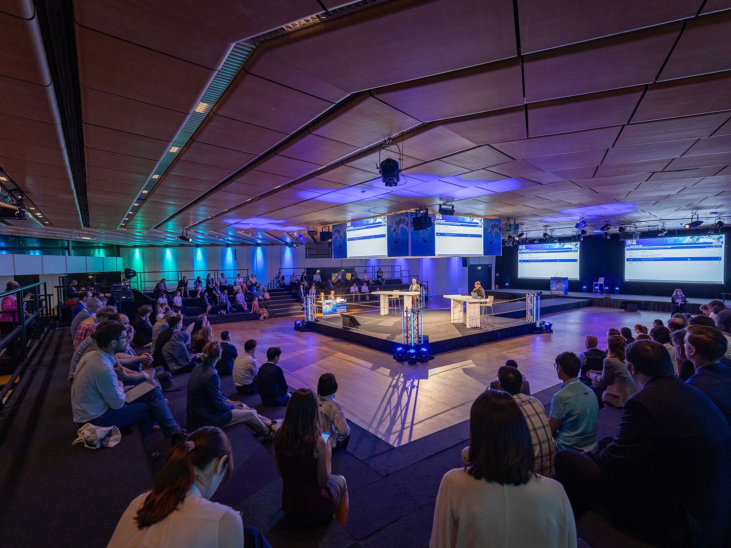 Foto: hell beleuchteter Podiumsdiskussions-Bereich mit einigen Panel-Teilnehmern vor einem Publikum