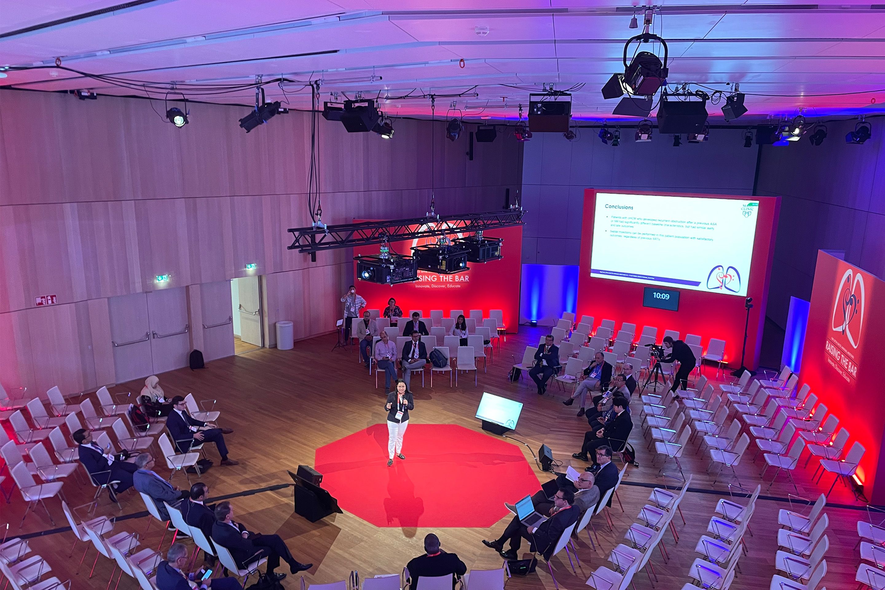 Foto: Raum mit Vortragendem im Zentrum, kreisfoermigen Sesselreihen mit Menschen und Leinwand