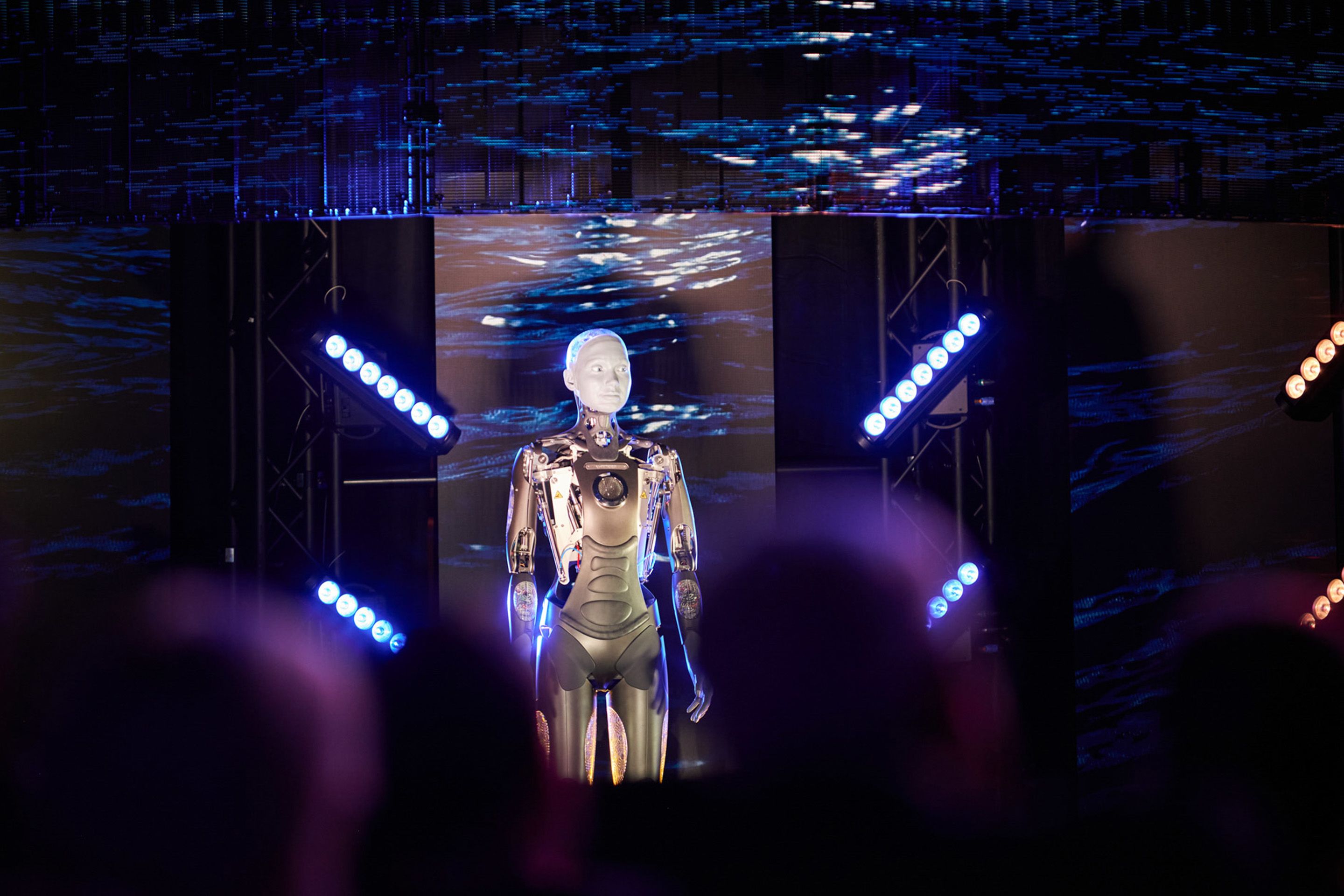 Foto: Roboter vor dunklem Hintergrund. Im Vordergrund ist schemenhaft ein Publikum zu sehen.