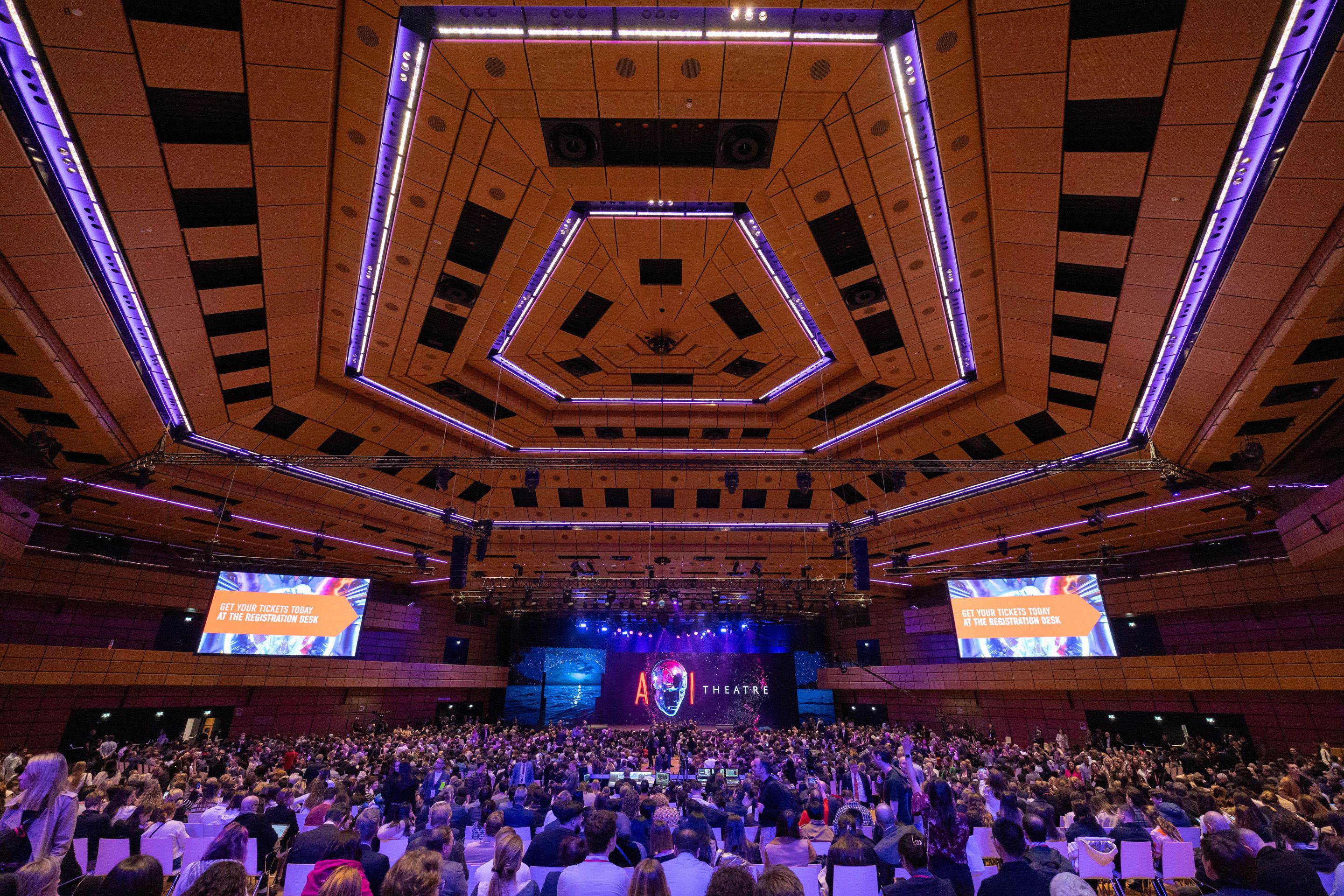Foto: dezent beleuchteter Saal voller Menschen, im Hintergrund eine Buehne und LED Waende