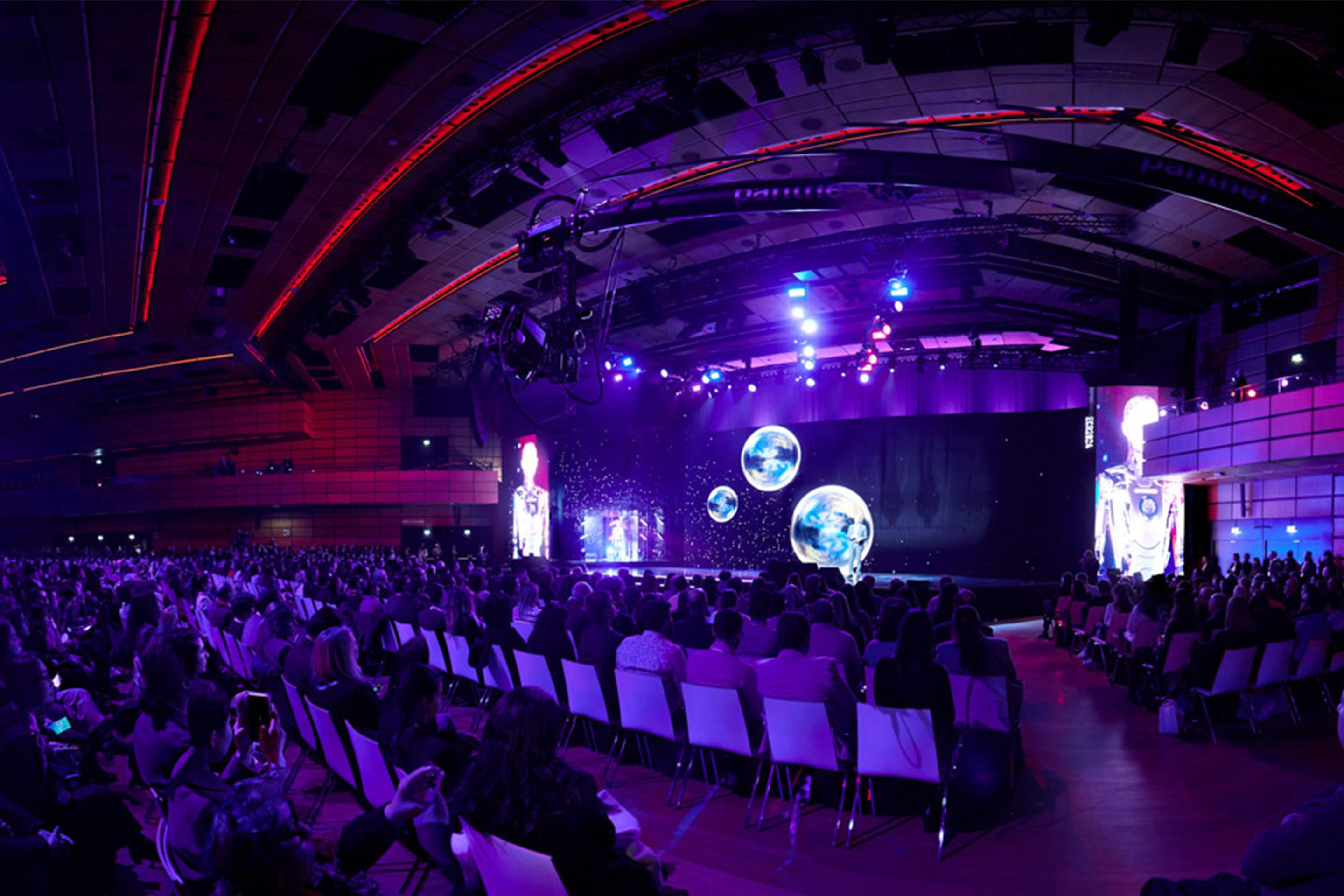 Dunkler Saal mit sitzenden Menschen, die auf eine Buehne schauen, auf der ein Moderator spricht