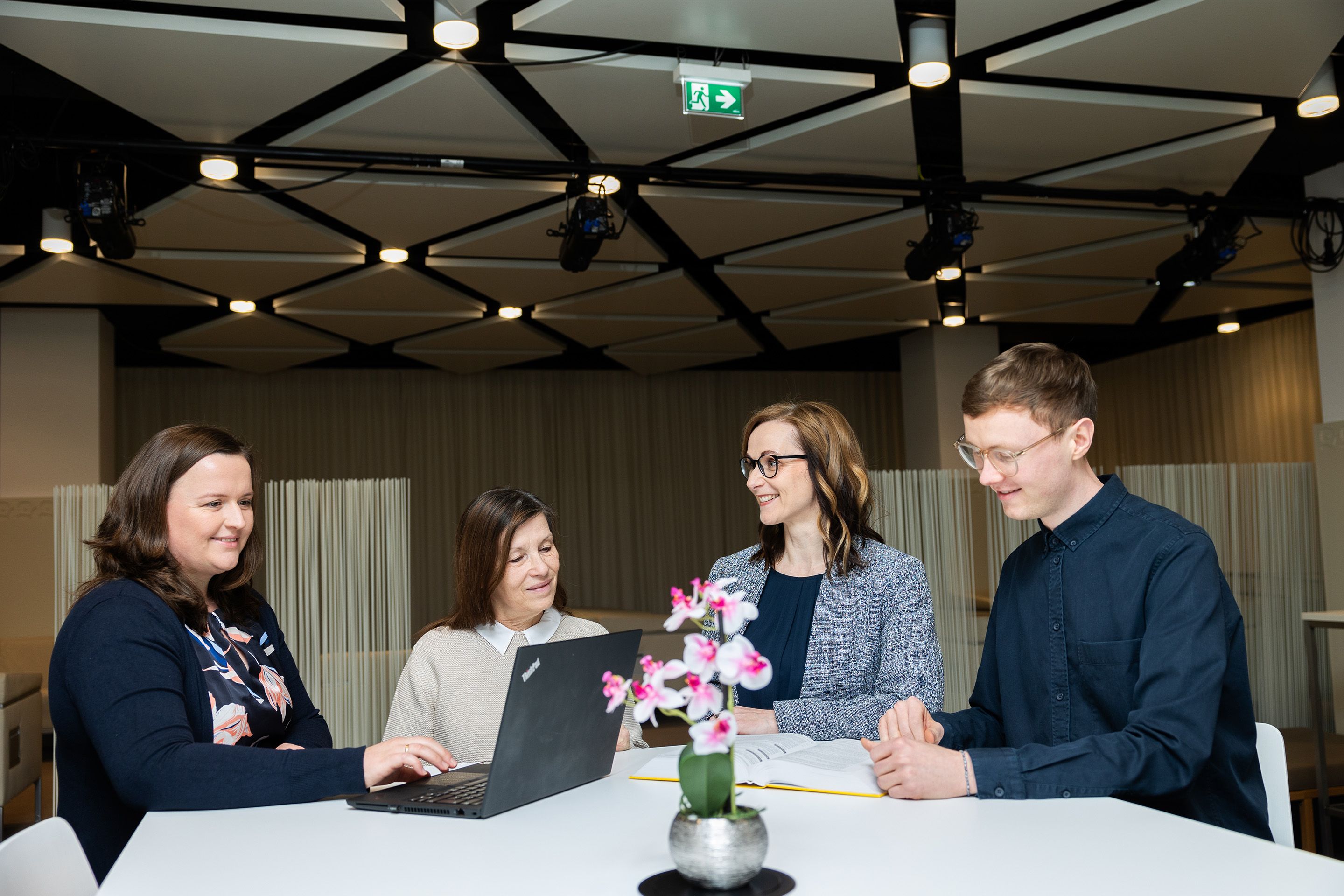 Drei Frauen und ein Mann stehen an einem Tisch mit einem Laptop fuer ein Meeting