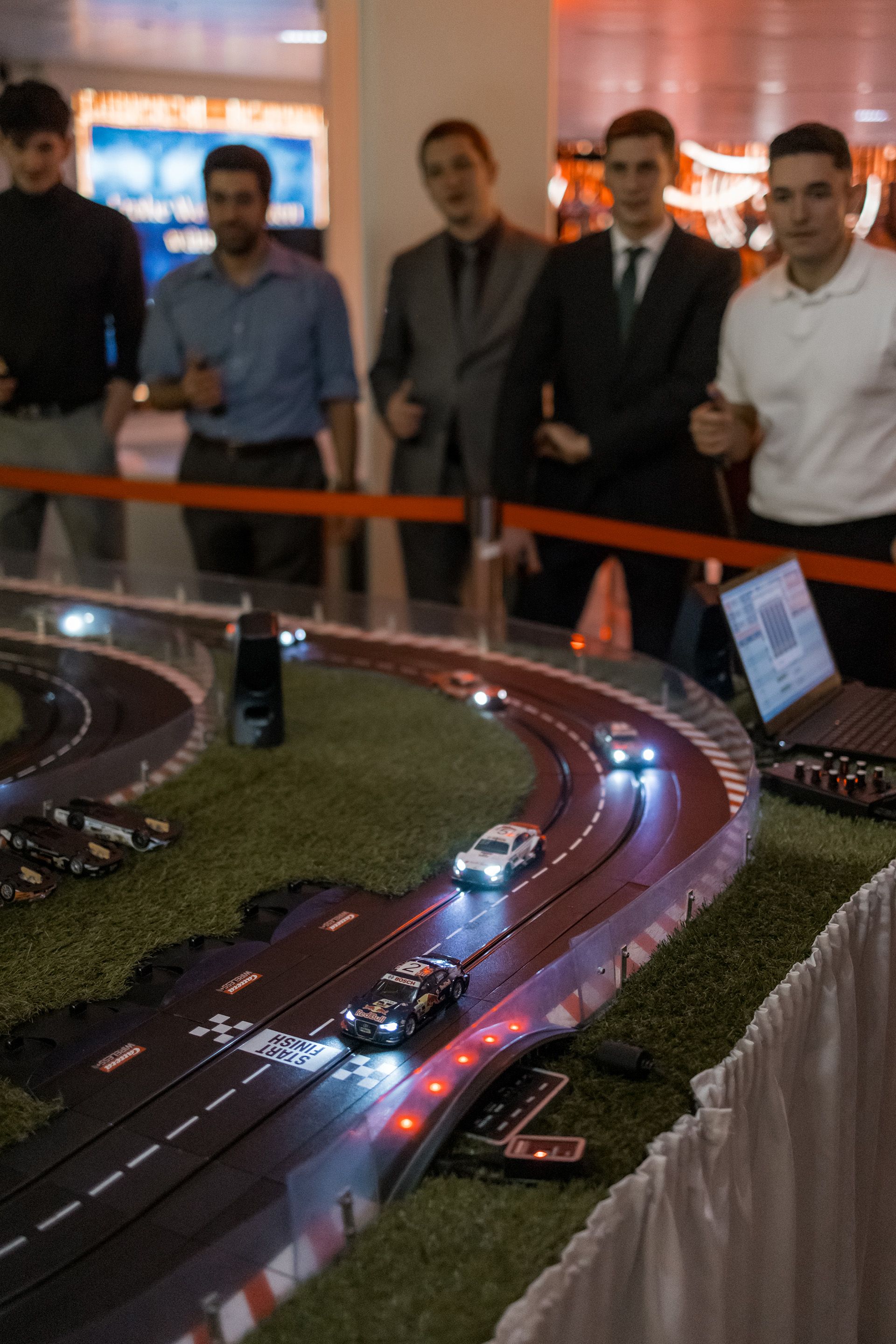 Eine Gruppe Maenner steht mit Joysticks vor einer Carrera-Bahn, auf der kleine Autos auf einer Rennstrecke fahren.