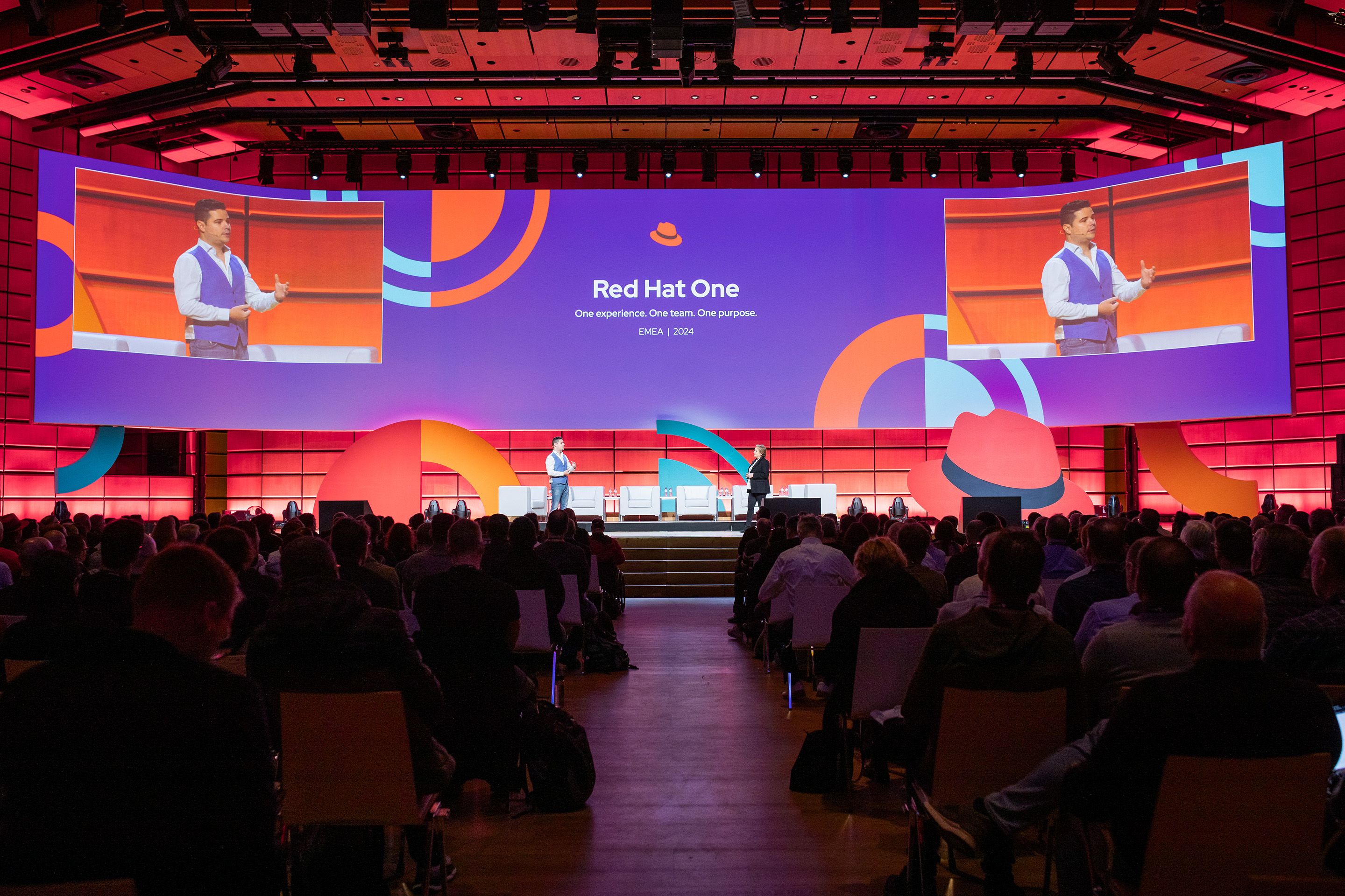 Rot beleuchtete Buehne, auf der 2 Personen stehen. Viele Reihen an Zuseher:innen blicken auf eine violett beleuchtete Leinwand.