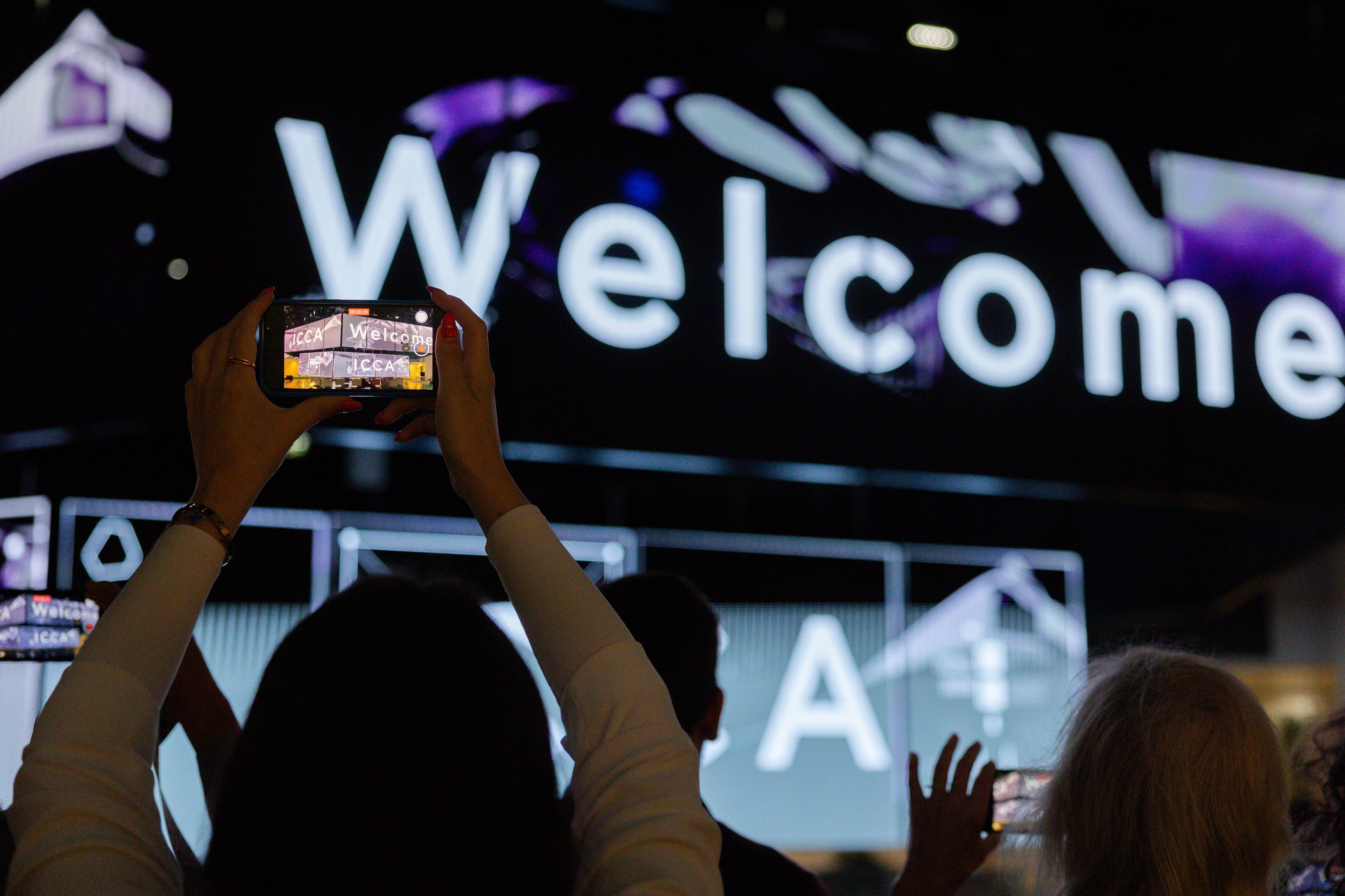 Frau filmt am Handy LED Wand im Austria Center Vienna mit Willkommensgruss fuer Teilnehmerinnen des ICCA Client Supplier European Venue Workshops 2024