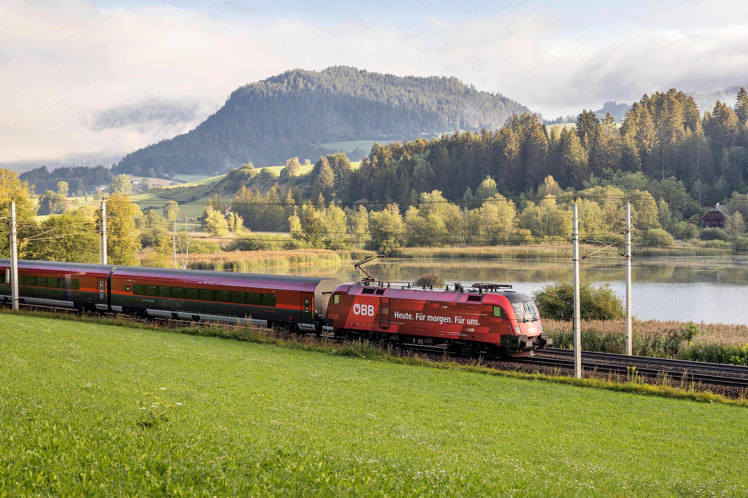 Roter OEBB Railjet faehrt durch Landschaft, im Hintergrund ein See und Berge.