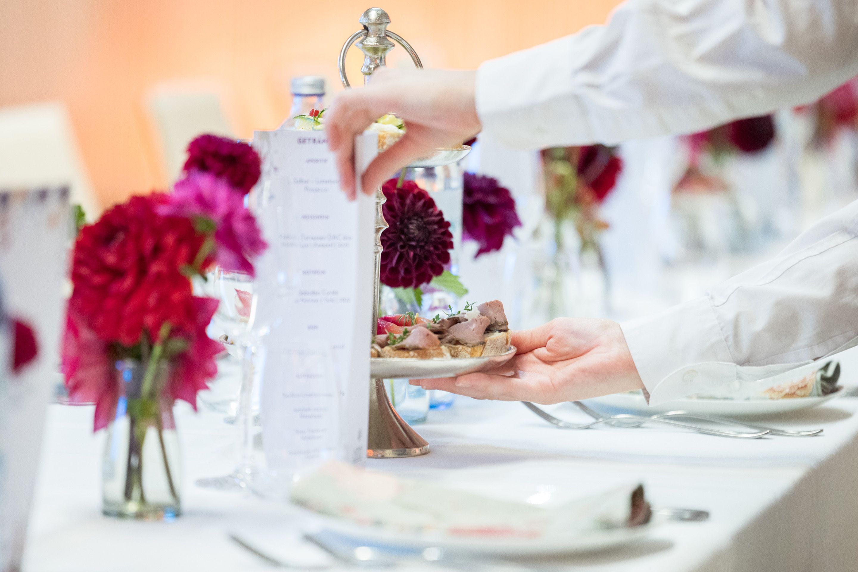Eine Person stellt einen Teller Essen auf einen weiss gedeckten Tisch mit roten Blumen