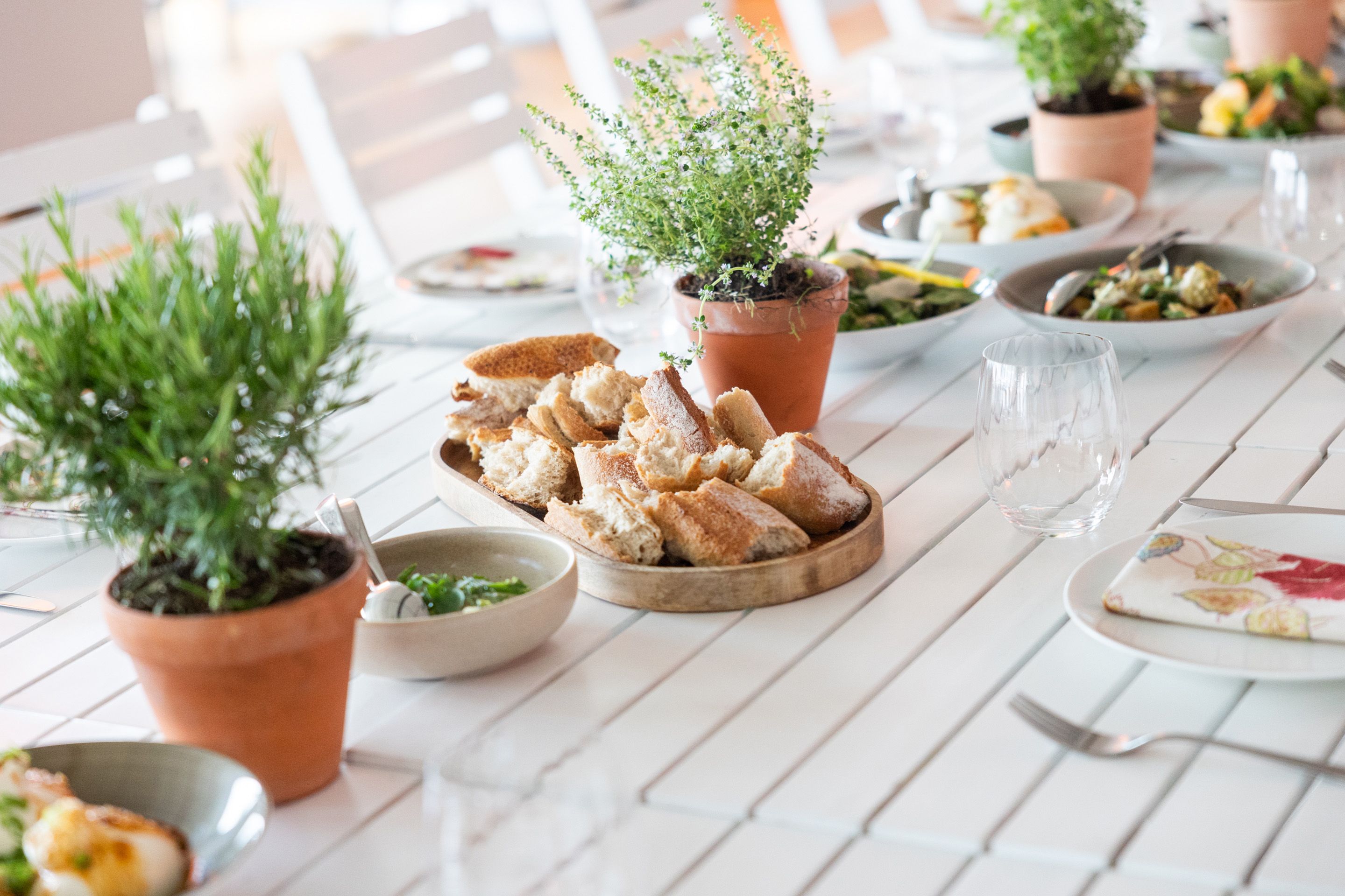 Weisser Tisch gedeckt mit Brot, Speisen und Kraeutertoepfen