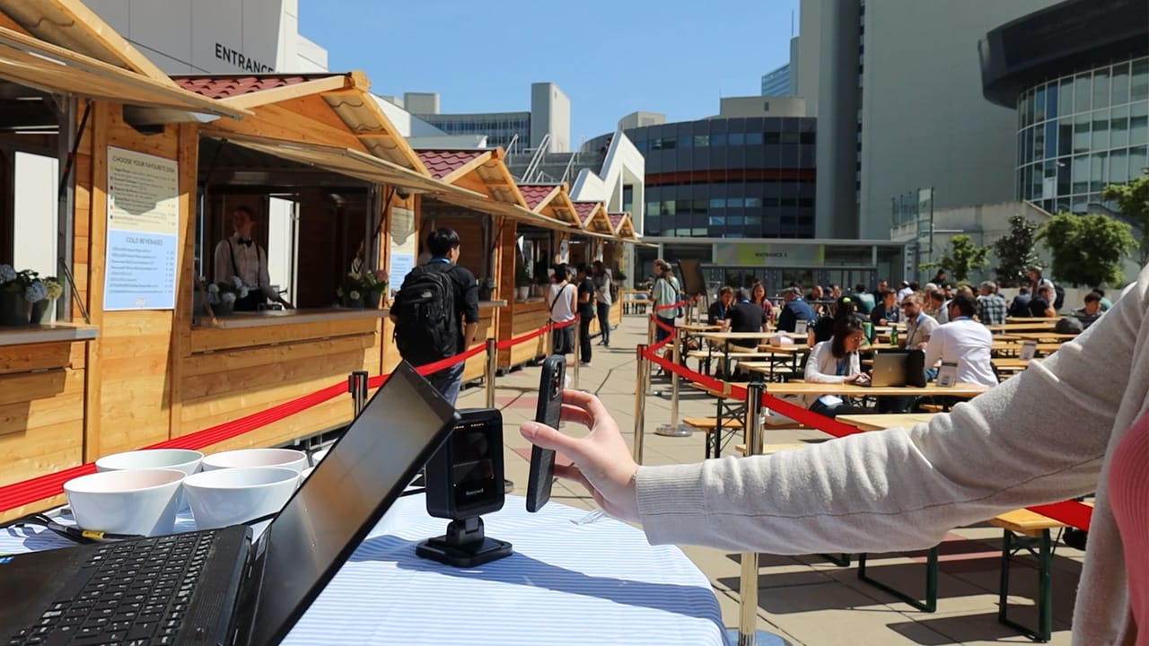 Eine Frau haelt ihr Smartphone an einen digitalen Reader, im Hintergrund Sitzbaenke und Holzstaende zur Essensausgabe am Vorplatz des Austria Center Vienna