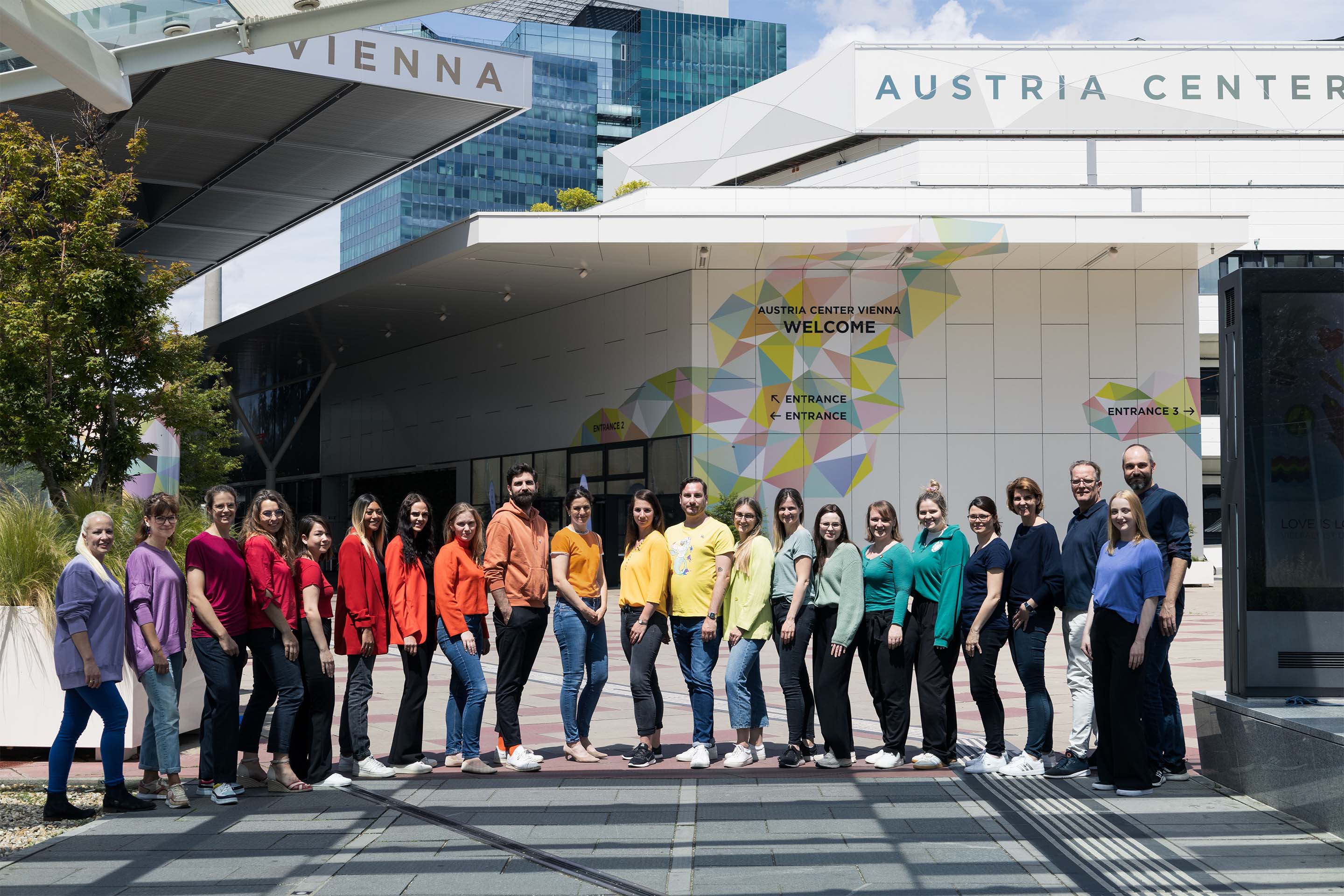 Mitarbeiterinnen des Austria Center Vienna in verschiedenfarbigen T-Shirts stehen in einer Reihe vor dem Austria Center Vienna