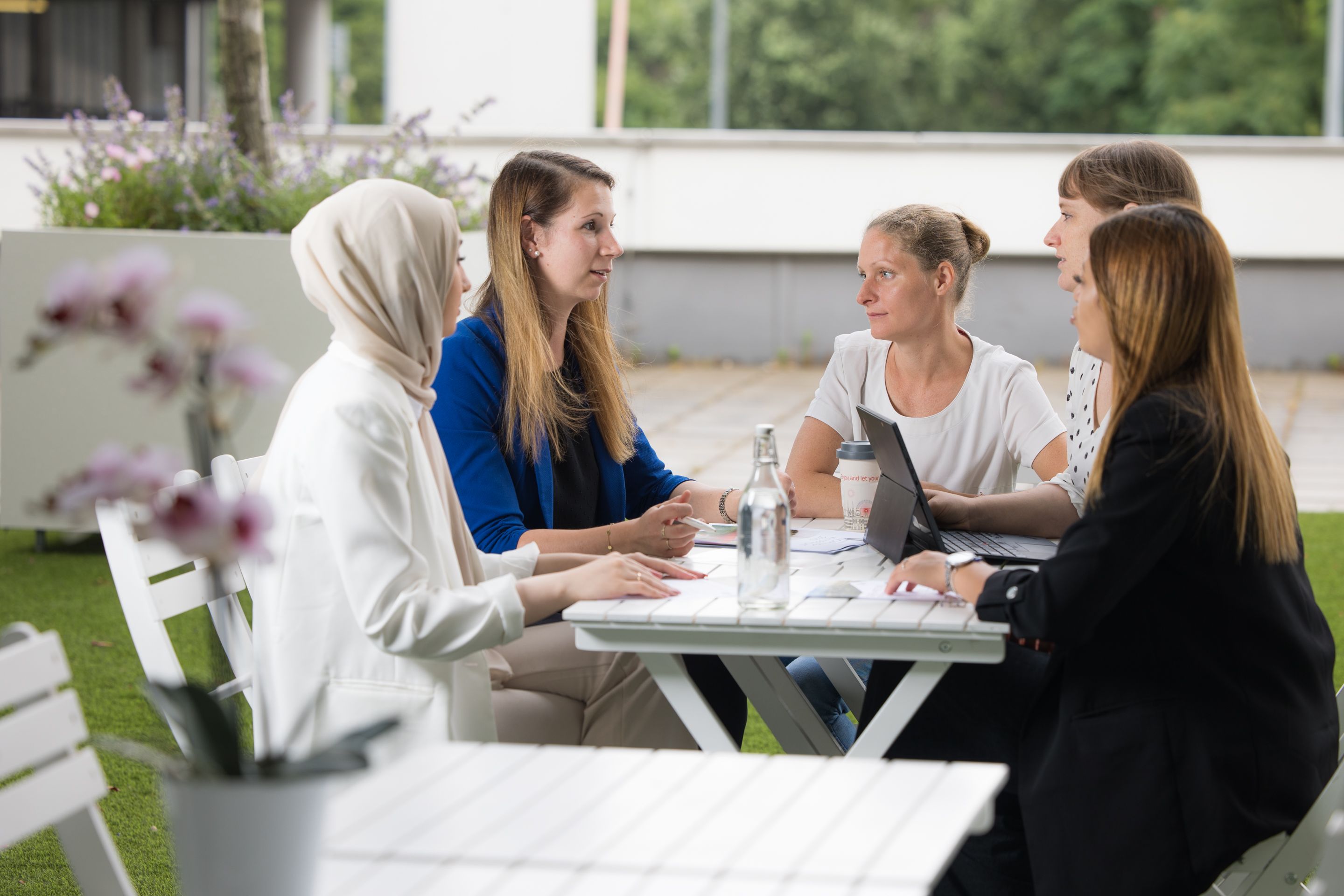Fünf Frauen, eine mit Kopftuch, im Gespräch am Tisch im Freien.